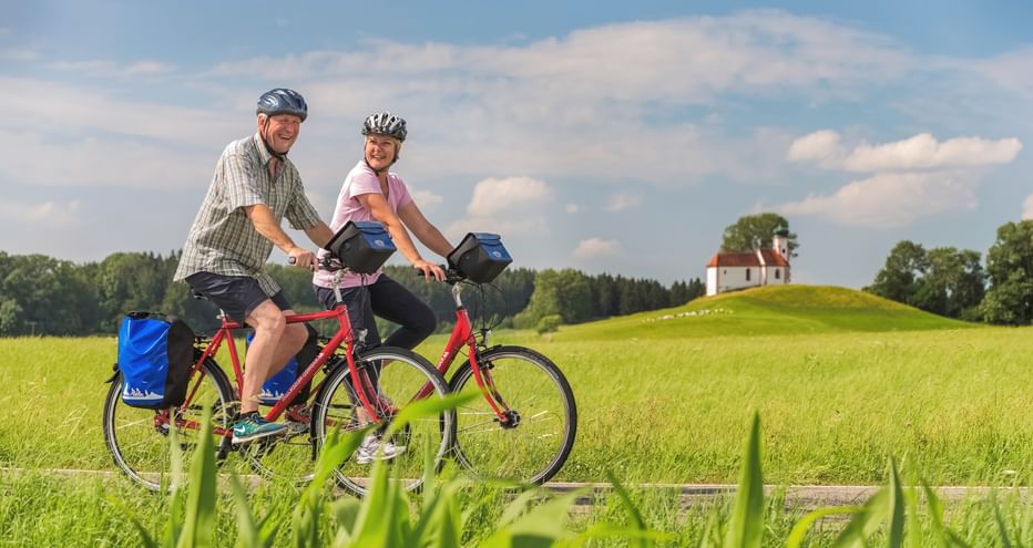 Zwei Radfahrer mit Packtaschen fahren durch grüne Felder in Bodensee-Oberschwaben. Eine kleine Kapelle auf einem Hügel und Wald im Hintergrund unter blauem Himmel.