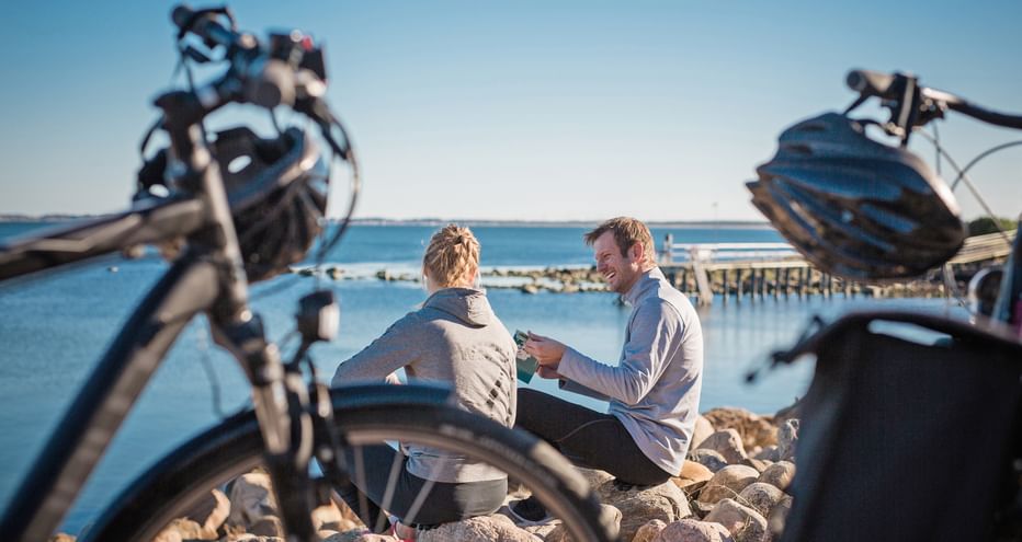 Zwei Radfahrer mit Rädern rasten auf felsigem Ufer am ruhigen blauen Wasser an Schwedens Westküste. Alte Holzpfähle im Hintergrund.