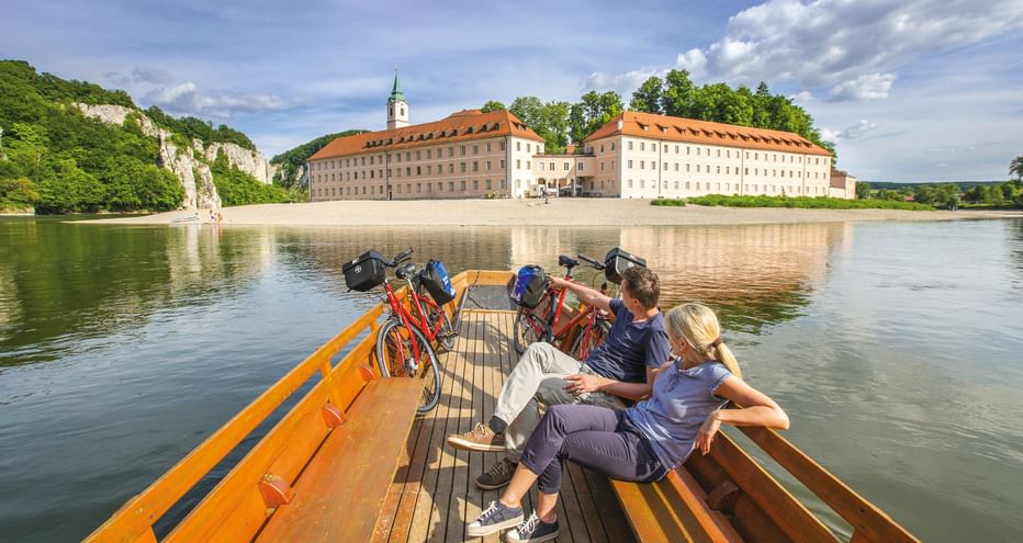Two cyclists with bikes on wooden ferry crossing the Danube River. Historic monastery with orange roof and green tower visible on riverbank.