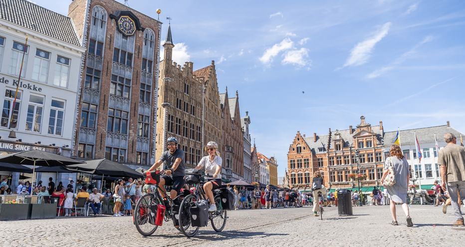 Zwei Radfahrer fahren über einen Kopfsteinpflasterplatz in Brügge, umgeben von historischen flämischen Gebäuden mit Treppengiebeln und Glockenturm.