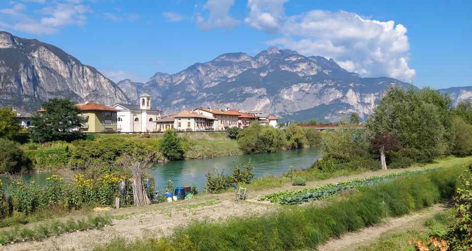Dorf mit Kirchturm neben türkisfarbener Etsch, umgeben von dramatischen Alpenbergen unter blauem Himmel mit weißen Wolken.