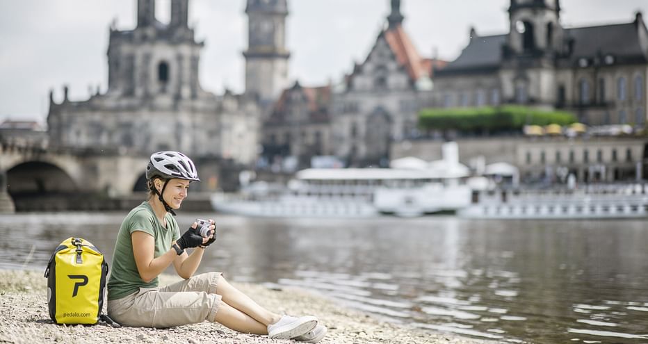 Radfahrerin mit Helm sitzt am Flussufer mit gelbem Rucksack und hält eine Kamera. Dresdner Altstadtpanorama mit Kirchen an der Elbe.